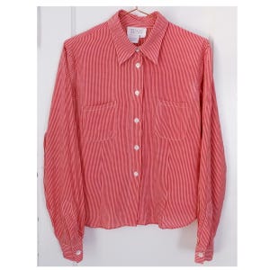 May include: A red and white striped long-sleeved button-down shirt with a collared neckline and two chest pockets. The shirt has a label that reads "TESS".