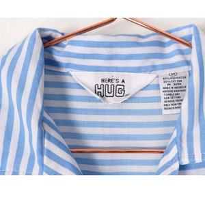 May include: Blue and white striped shirt with a label that reads "HERE'S A HUG" and care instructions. The shirt is size M.