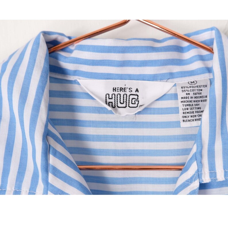 May include: Blue and white striped shirt with a label that reads "HERE'S A HUG" and care instructions. The shirt is size M.