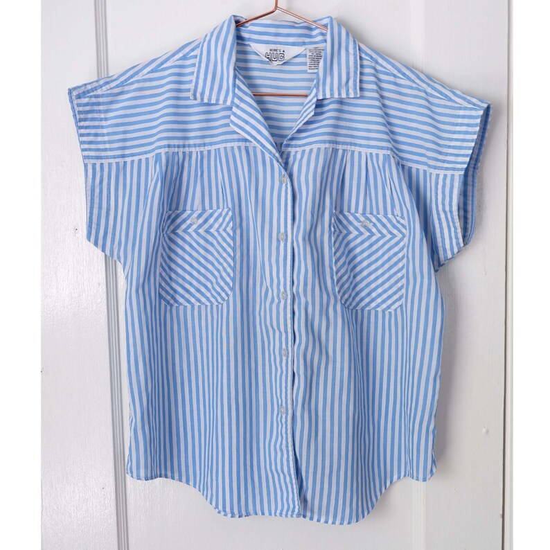 May include: A blue and white striped button-down shirt with short sleeves and two chest pockets. The shirt has a collared neckline and a button closure. The brand label reads "Kurt A.  Albus".