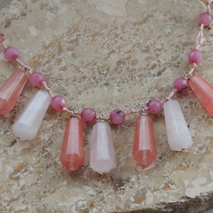 May include: A necklace featuring a light pink cord strung with alternating round pink beads and faceted, elongated, translucent pink and pale white stone pendants. The pendants hang vertically from the cord in a repeating pattern of colors.