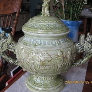 May include: A large green ceramic serving bowl with a lid. The bowl is decorated with raised floral and figural designs. The lid has a small finial in the shape of a bird. The bowl has two handles shaped like dragons.
