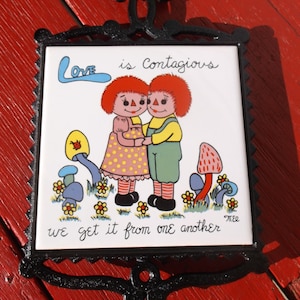 May include: A decorative black metal wall hanging with a ceramic tile featuring a cartoon couple. The tile reads "Love is Contagious, we get it from one another." The couple is hugging, surrounded by flowers and mushrooms.