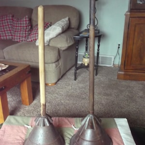 Op de afbeelding: Twee antieke ontstoppers. Elk heeft een houten handvat en een metalen beker. Eén handvat is licht van kleur, de andere donkerder. De metalen bekers zijn conisch en vertonen tekenen van slijtage.