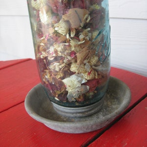 Może przedstawiać: Szklany słoik wypełniony suszonymi kwiatami i płatkami, w tym różowymi, czerwonymi i białymi, stoi na szarej metalowej podstawie. Podstawa znajduje się na czerwonej drewnianej powierzchni.
