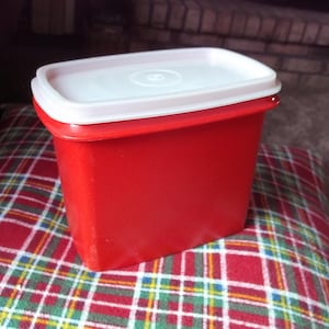 May include: A red rectangular food storage container with a white lid. The container is sitting on a plaid pillow. The lid has a circular design in the center.