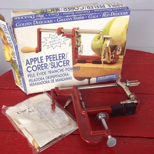 May include: An apple peeler/corer/slicer with a red metal frame and a wooden handle, next to its original box. The box features images of apples and text in multiple languages, including "Golden Delicious" and "Granny Smith."