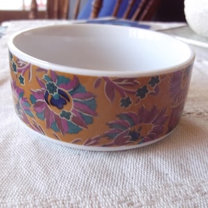 Può includere: Una ciotola in ceramica bianca con un motivo floreale. L'esterno della ciotola presenta uno sfondo color senape adornato con motivi floreali viola, verde acqua e blu. La ciotola è appoggiata su una superficie bianca e strutturata.