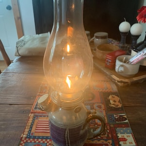 May include: A lit oil lamp with a clear glass chimney sits on a colorful patterned table runner. The lamp has a ceramic base with a handle and a purple band. The flame glows brightly, casting a warm light.