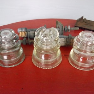 May include: Three vintage glass insulators with different designs. The insulators are clear glass and have a slightly yellowed tint. They are sitting on a red surface.