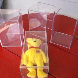 May include: Clear acrylic display cases in various shapes and sizes. One case contains a yellow teddy bear with a red bow. The cases are on a red wooden surface.