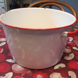 Może przedstawiać: Duży, zabytkowy garnek emaliowany z białym wnętrzem i bladoróżowym zewnętrzem. Obręcz i uchwyty są czerwone. Garnek stoi na czerwonym obrusie z wzorem jabłek. Prawdopodobnie służy do gotowania lub przechowywania.