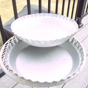 May include: Two white metal bird baths with scalloped edges. The top bath is slightly elevated above the lower one, creating a tiered effect. The baths are outdoors on a deck.