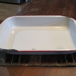 May include: White enamel baking dish with a red rim. The dish is rectangular and has a slight curve to the sides.