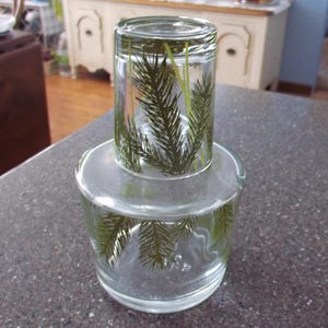 May include: Two clear glass tumblers, one inverted atop the other. Both glasses feature a green fern pattern. The glasses are sitting on a speckled gray countertop. The image is well-lit.