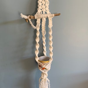 Könnte beinhalten: Ein weißer Makramee-Pflanzenhänger mit einem kleinen weißen Keramiktopf, der daran hängt. Der Hänger ist an einem Treibholzast befestigt.