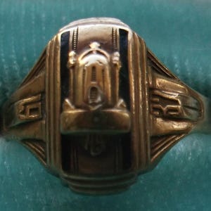 May include: A vintage gold-coloured ring with a central architectural design, possibly a school or organisation emblem. The ring features black accents and intricate detailing, with a rectangular top and a wide band. The ring is set against a teal background.