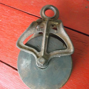 Op de afbeelding: Een antieke katrol met een metalen frame en een donker, cirkelvormig wiel. De katrol heeft een lus aan de bovenkant om op te hangen en staat op een rode houten ondergrond. Het woord "CRANE" is zichtbaar op het wiel.