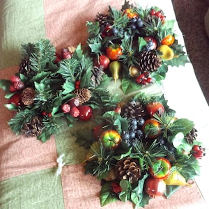 May include: Three decorative holiday wreaths featuring pine needles, pine cones, red berries, and various fruits. The wreaths are arranged on a patterned surface, creating a festive and seasonal display.
