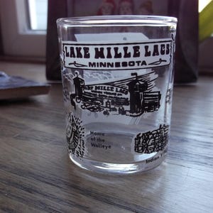 May include: Clear glass tumbler with black and white graphics. The text "Lake Mille Lacs Minnesota" is at the top, with illustrations of buildings and the text "Home of the Walleye" and "Indian Village" below.