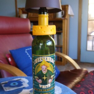 May include: A green wine bottle with a yellow cheese-shaped bottle stopper. The bottle label says "Cheesehead White Wine for the Rest of Us" and features a cartoon character wearing a green shirt and hat. The bottle is under a brown lampshade.