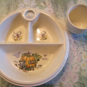 May include: A white divided plate with a handle, featuring a pumpkin house and two cartoon animals. The plate is on a floral tablecloth, next to a white cup.