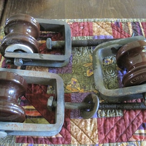 May include: Four brown ceramic insulators with metal clamps. The insulators have a rounded shape and are held in place by the clamps, which have threaded rods and nuts for adjustment. The items are on a colorful patchwork quilt.