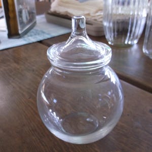 May include: A clear glass jar with a round body and a lid. The jar has a small handle on top of the lid. The jar is sitting on a wooden surface. Other glass jars are in the background.
