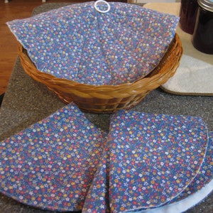 May include: A wicker basket lined with a blue and white floral fabric. The basket is sitting on a table with two additional pieces of the same fabric.