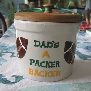 Op de afbeelding: Witte keramische pot met een houten deksel. De pot is versierd met groene en gele verf en heeft twee voetballen en de tekst "Dad's A Packer Backer".