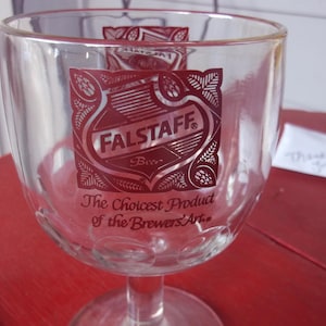 May include: Clear glass beer goblet with a red Falstaff Beer logo and text. The logo is a shield shape with the words "Falstaff Beer" and "The Choicest Product of the Brewers' Art." The goblet has a stem and a wide bowl.