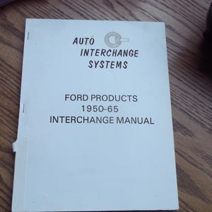 May include: A white manual titled "AUTO INTERCHANGE SYSTEMS" with the text "FORD PRODUCTS 1950-65 INTERCHANGE MANUAL" in black. The manual is on a wooden surface.