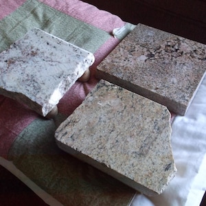 May include: Three granite coasters with a rough, natural edge. The coasters are various shades of brown, beige, and white with flecks of black. The coasters are on a pink and green patterned fabric.