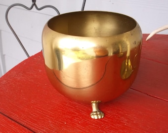 May include: A gold-colored, round bowl with a single paw-shaped foot. The bowl has a wide opening and a smooth, reflective surface. It sits on a red wooden surface. A decorative antler and a brass object are in the background.
