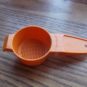 May include: An orange Tupperware strainer with a handle and a perforated bowl. The handle has a hole for hanging. The strainer is made of durable plastic and is designed for kitchen use.