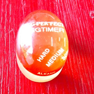 May include: An egg-shaped kitchen timer with a red and white design. The timer has the words "G-PERFECT EGGTIMER" printed on it, along with "HARD", "MEDIUM", and "SOFT" indicators. The base of the timer is a light cream color.