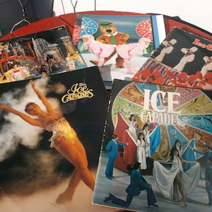 May include: A collection of vintage Ice Capades show programs. The covers feature colorful illustrations of ice skaters, cartoon characters, and performance scenes. The programs are arranged on a red surface.