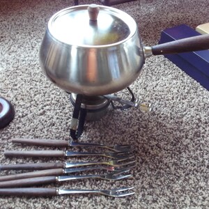 May include: A vintage fondue set with a stainless steel pot, wooden-handled forks, and a burner. The pot has a lid and a wooden handle. The set includes a stand and a set of fondue forks. The items are on a carpeted surface.