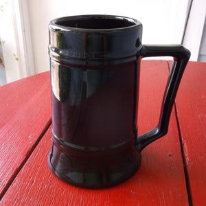 May include: A black ceramic beer stein with a handle, sitting on a red wooden surface. The stein has a cylindrical shape with a slightly flared base and a thick, angular handle. The glossy finish reflects the surrounding light.