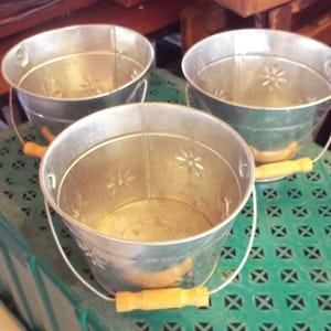 May include: Three galvanized metal buckets with white handles and wooden grips. Each bucket has a floral design and is sitting on a green plastic surface. The buckets are empty and ready to be used.