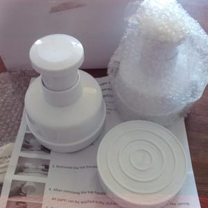 May include: White burger press components, including a press, a base, and a top handle, are displayed on a wooden surface. One component is wrapped in bubble wrap. Instructions are partially visible.