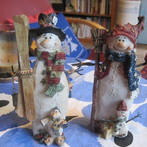 May include: Two snowman figurines, one holding skis and the other a broom. The snowman with skis has a red and green scarf and a green hat. The snowman with the broom has a blue scarf and a red hat. Both snowmen have white faces and are decorated with snowflakes.
