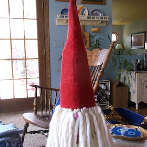May include: A tall, red felt gnome with a long, pointed hat and a white, shaggy beard. The gnome has a pink heart-shaped nose and is a festive holiday decoration. The gnome is approximately 24 inches tall.