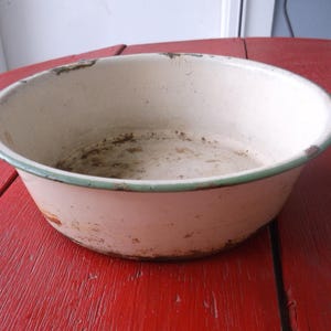 May include: An antique enamel bowl, off-white with a pale green rim, showing signs of age and wear. The bowl has some rust and discolouration. It is placed on a red wooden surface.