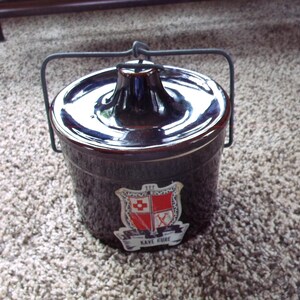 May include: A dark brown ceramic crock with a lid and a metal handle. The crock has a decorative emblem with red and white details and the text "Kave Kure". This vintage container is likely used for food storage.