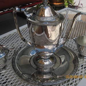 Könnte beinhalten: Ein silbernes Teeservice, bestehend aus einer Teekanne, einer Zuckerdose, einem Sahnekännchen und einem Serviertablett. Das Set hat aufwendige Details und eine reflektierende Oberfläche. Die Teekanne hat einen Deckel und einen gebogenen Ausguss. Das Set wird auf einer weißen Metalloberfläche präsentiert.