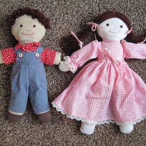 May include: Two handmade cloth dolls. One doll is dressed in blue dungarees and a red heart-patterned shirt. The other doll wears a pink gingham dress with white lace trim. Both dolls have brown yarn hair and smiling faces.