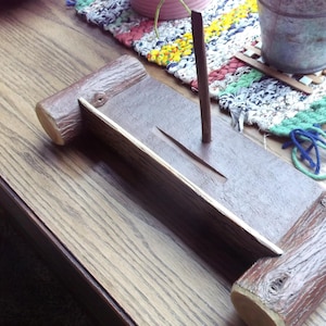 May include: A wooden incense holder with a brown finish. The holder features a rectangular base with two cylindrical log-like supports on each end. A vertical stick and a thin, angled piece are in the center.