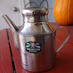 May include: A silver stainless steel oil and vinegar cruet with a spout and a handle. The cruet has a sticker on the front that says "XinXierJingPin" and "新希尔精品".
