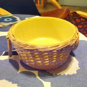 May include: A yellow bowl with a wicker basket exterior. The bowl has two handles on the sides. The bowl is sitting on a blue and white patterned tablecloth.